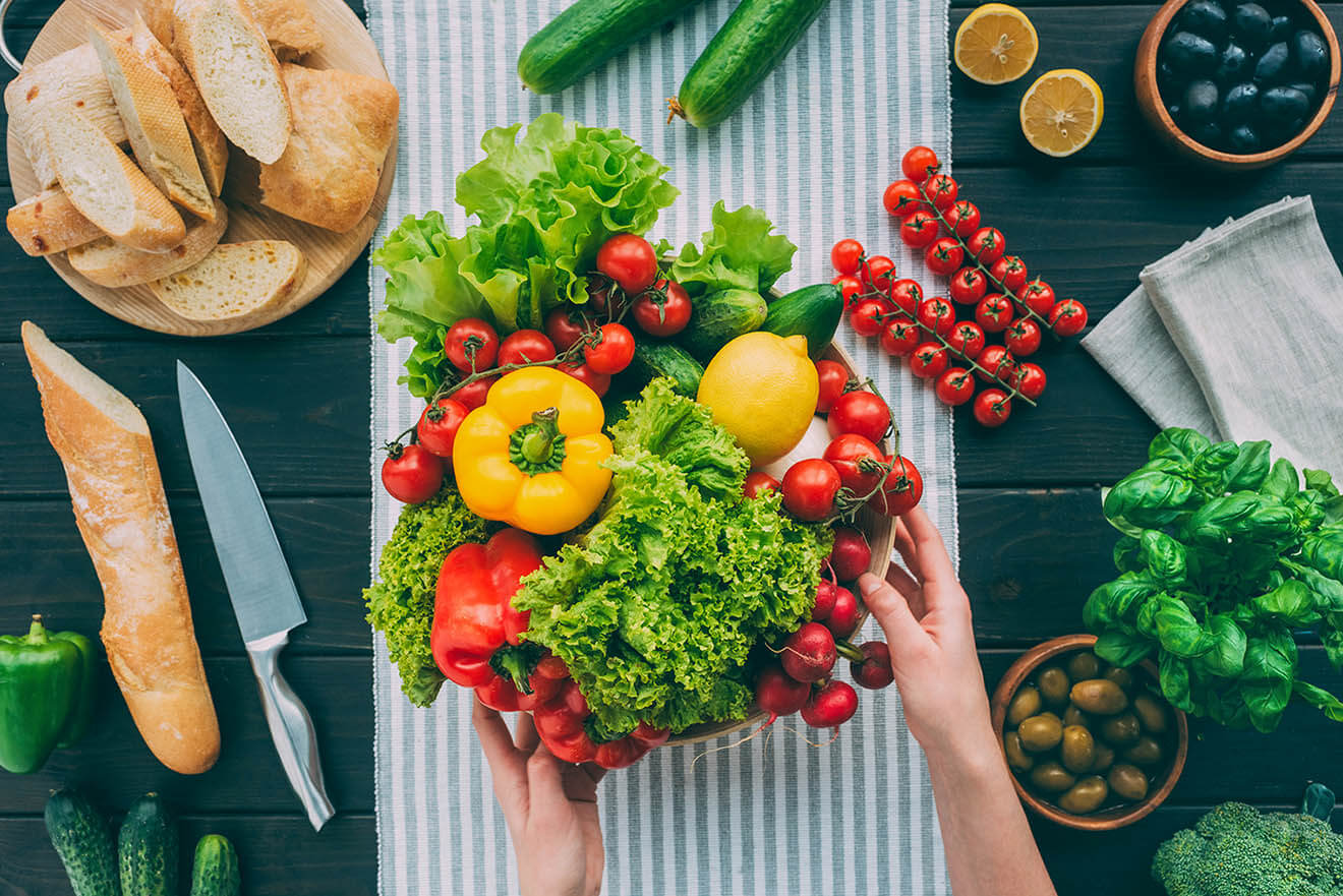 Mãos sobre mesa com legumes, verduras e frutas frescas, fontes de fibra
